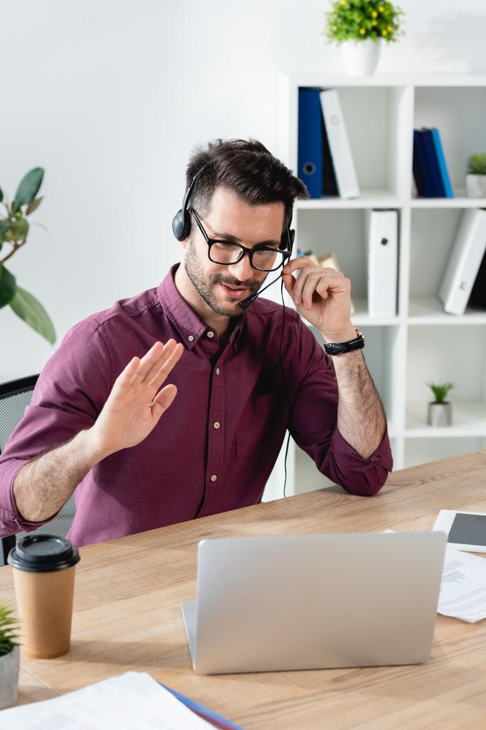 glimlachende zakenman in headset met hallo gebaar tijdens videochat op laptop - Foto, afbeelding