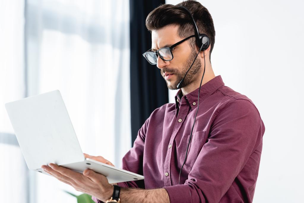 young, serious businessman in headset holding laptop during video call on laptop - Photo, Image