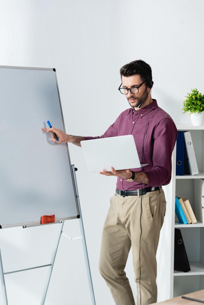 συμπυκνωμένος επιχειρηματίας κρατώντας φορητό υπολογιστή, ενώ στέκεται κοντά flipchart - Φωτογραφία, εικόνα