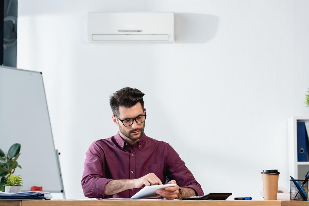 hombre de negocios guapo usando tableta digital mientras está sentado en el lugar de trabajo bajo el aire acondicionado en la pared
 - Foto, imagen