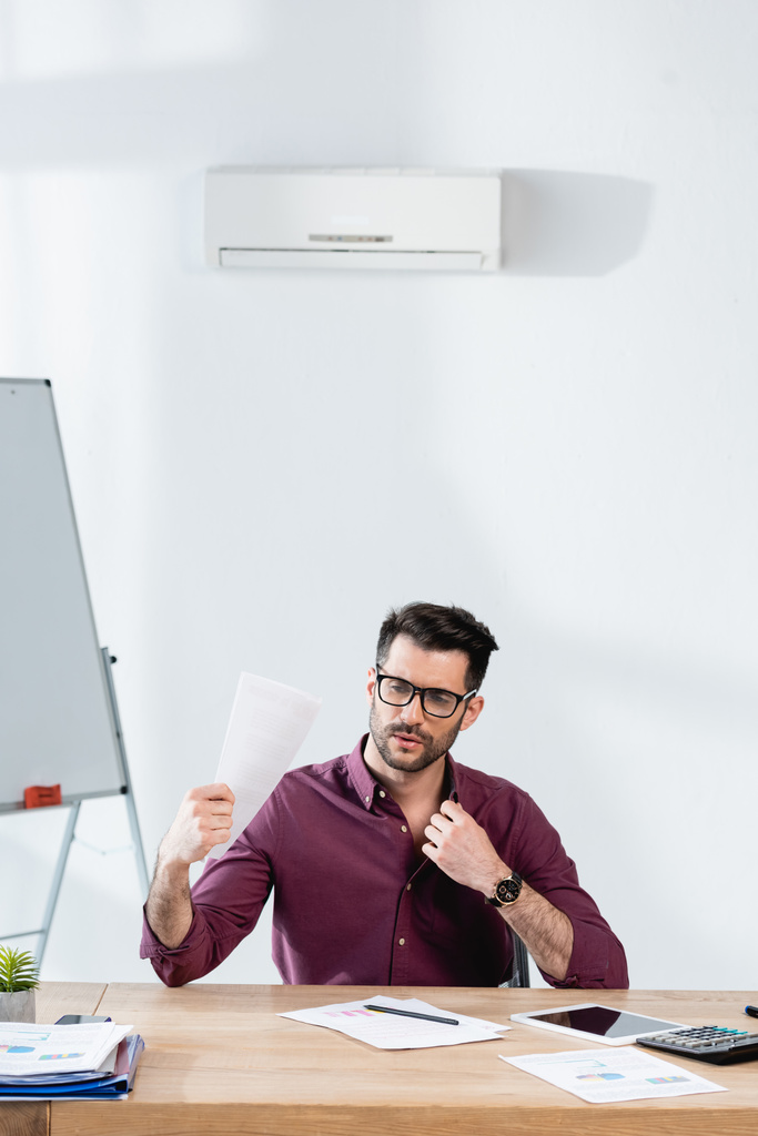 joven hombre de negocios tocando camisa y agitando papel mientras sufre de calor en la oficina
 - Foto, Imagen