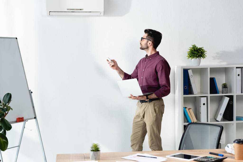 νεαρός επιχειρηματίας κρατώντας φορητό υπολογιστή και ενεργοποίηση κλιματιστικού με τηλεχειριστήριο - Φωτογραφία, εικόνα