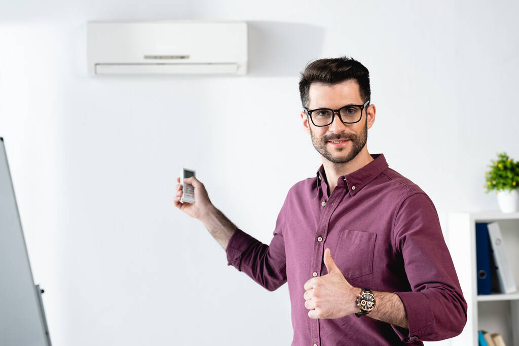 smiling businessman showing thumb up while switching on air conditioner with remote controller - Photo, Image