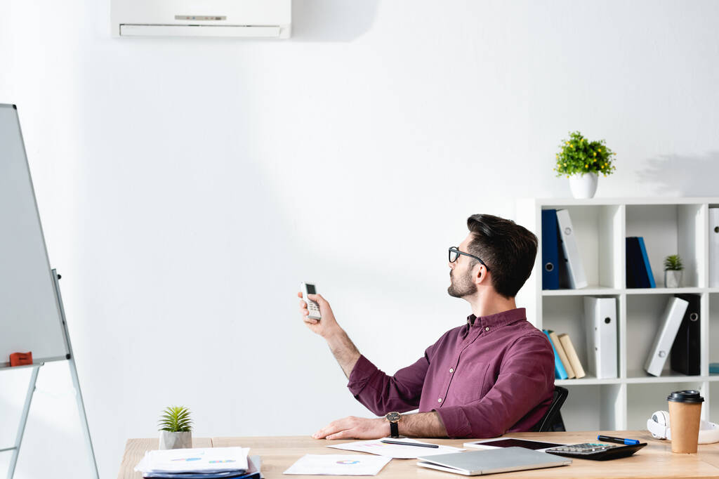jeune homme d'affaires assis sur le lieu de travail et allumant le climatiseur avec télécommande - Photo, image