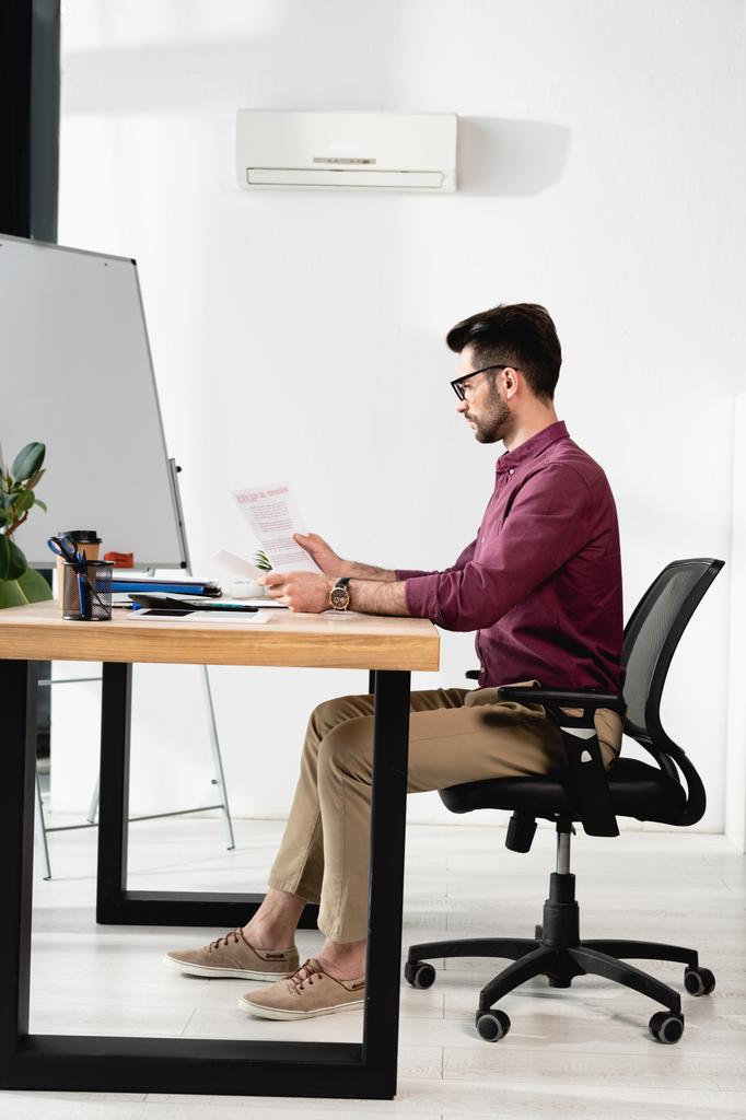 widok z boku biznesmen patrząc na papiery podczas siedzenia w miejscu pracy w pobliżu klimatyzatora  - Zdjęcie, obraz