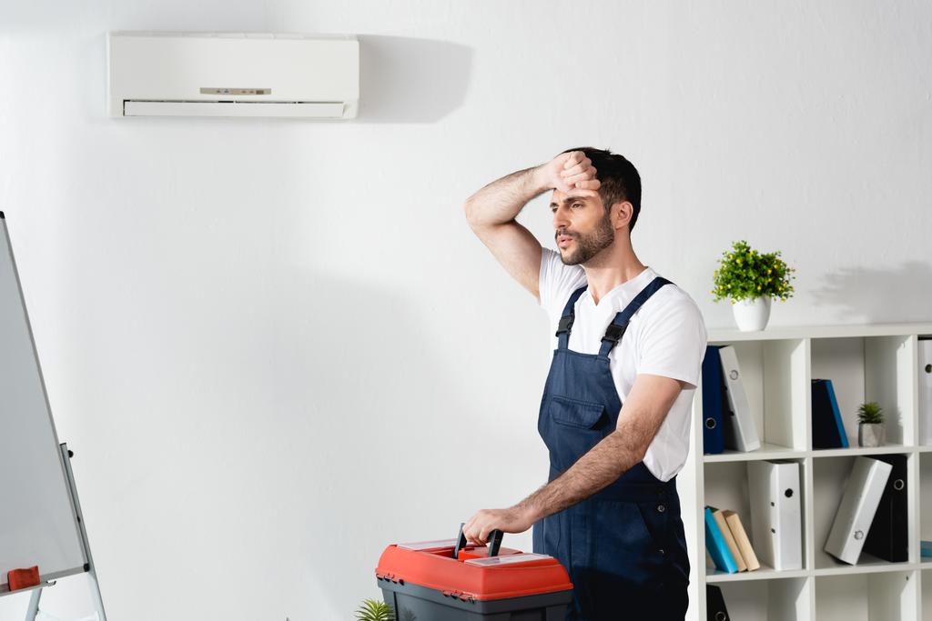 Arbeiter berührt Stirn, während er mit Werkzeugkiste in der Nähe einer defekten Klimaanlage steht und unter Hitze leidet - Foto, Bild