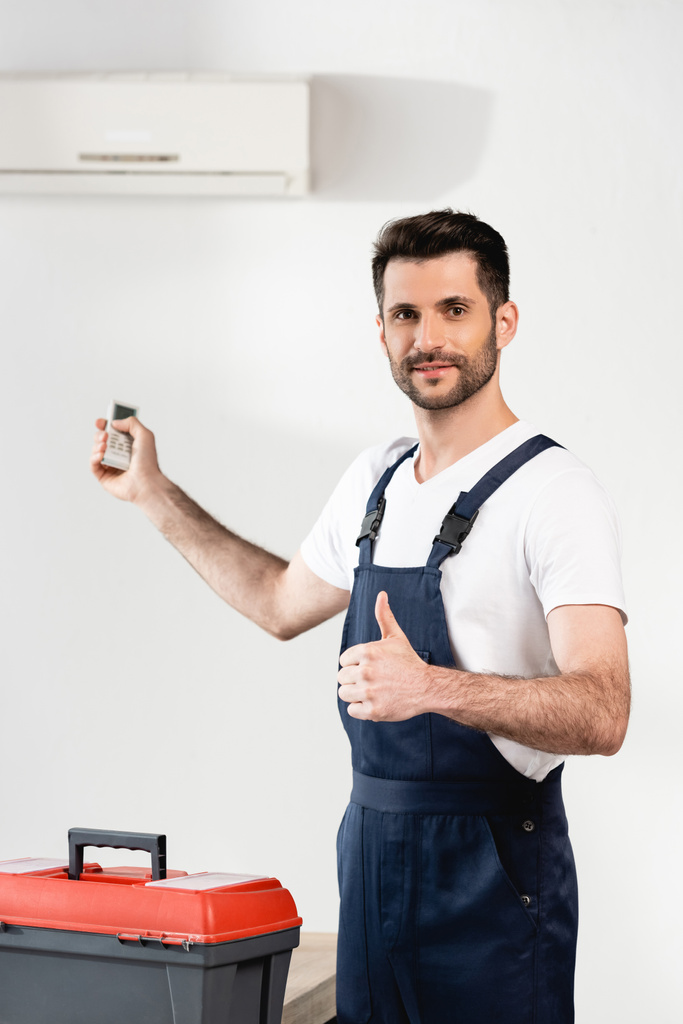 glimlachende reparateur met afstandsbediening en duim in de buurt van gereedschapskist en airconditioner op de muur - Foto, afbeelding