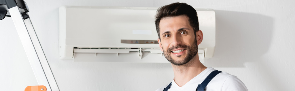 panoramic crop of happy repairman smiling at camera near air conditioner on wall - Photo, Image