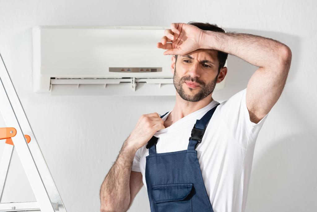 reparador insatisfecho tocando la frente mientras está de pie cerca del acondicionador de aire roto y sufriendo de calor
 - Foto, Imagen