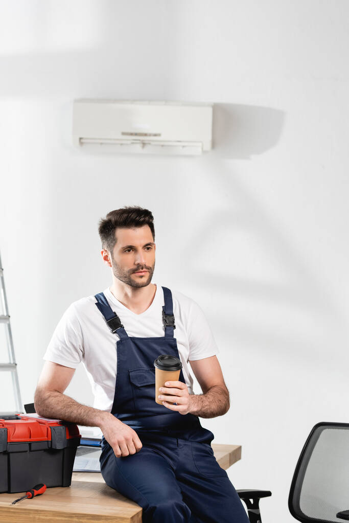bonito reparador segurando café para ir enquanto sentado na mesa perto de caixa de ferramentas sob ar condicionado na parede
 - Foto, Imagem