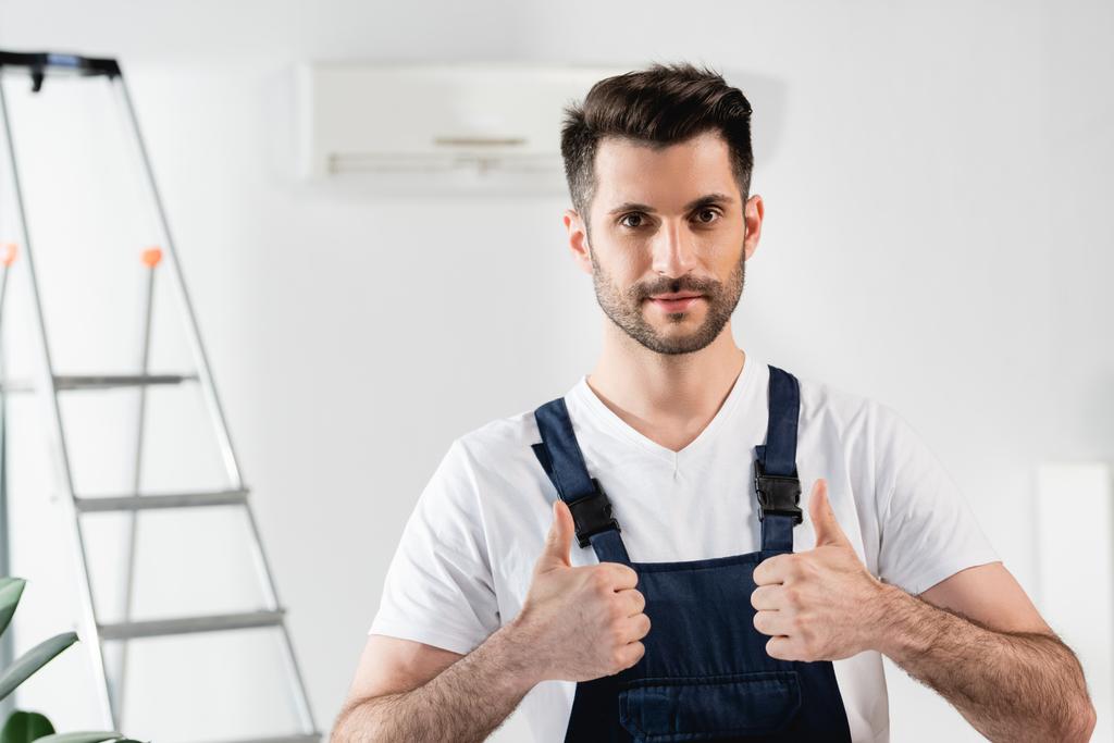 pohledný opravář ukazuje palce nahoru, zatímco stojí v blízkosti žebříku a klimatizace na stěně - Fotografie, Obrázek