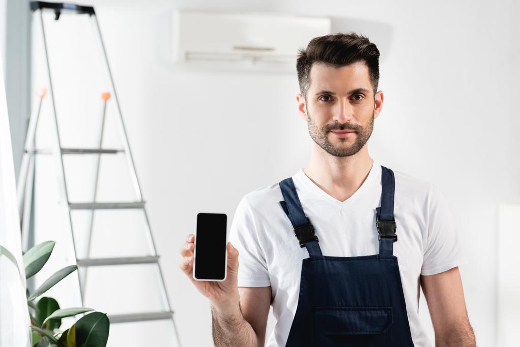 gutaussehender Reparateur zeigt Smartphone mit leerem Bildschirm neben Leiter und Klimaanlage an der Wand  - Foto, Bild