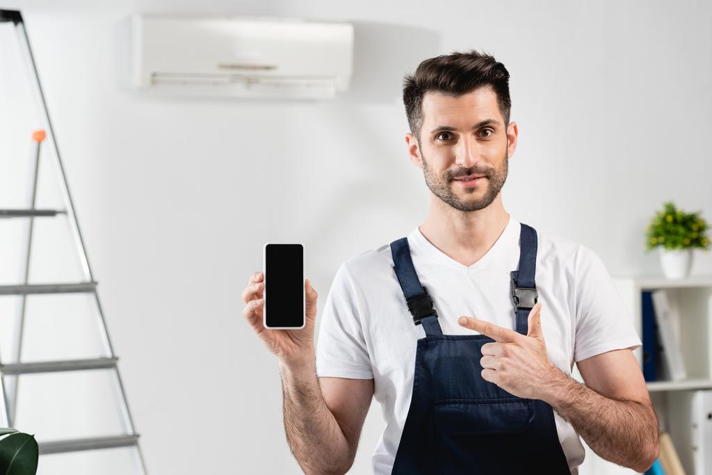 riparatore sorridente che punta a smartphone con schermo vuoto vicino al condizionatore d'aria a parete - Foto, immagini