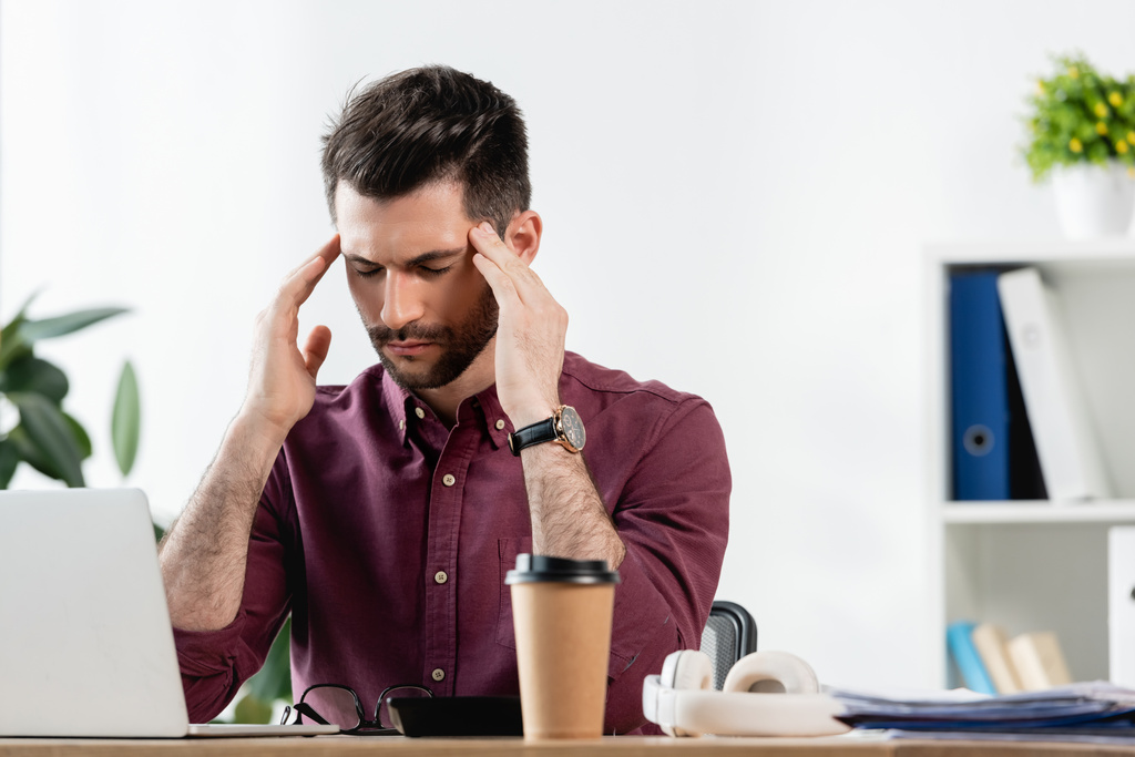 vyčerpaný podnikatel se zavřenýma očima dotýká hlavy, zatímco sedí v blízkosti notebooku a kávy jít - Fotografie, Obrázek