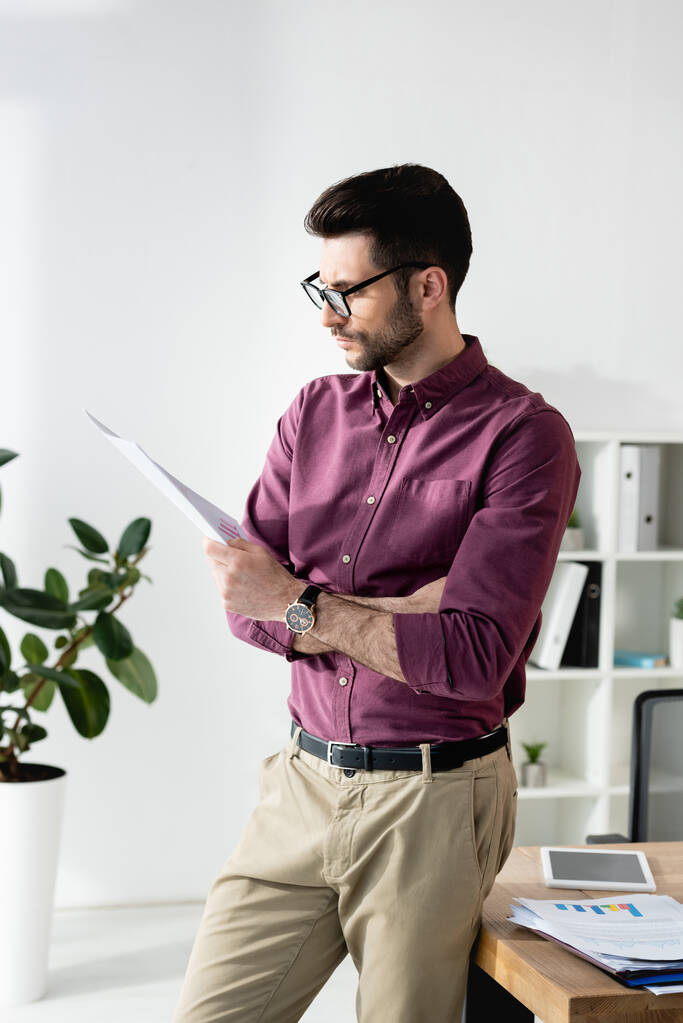 attentive businessman reading document while standing at desk near potted plant - Photo, Image