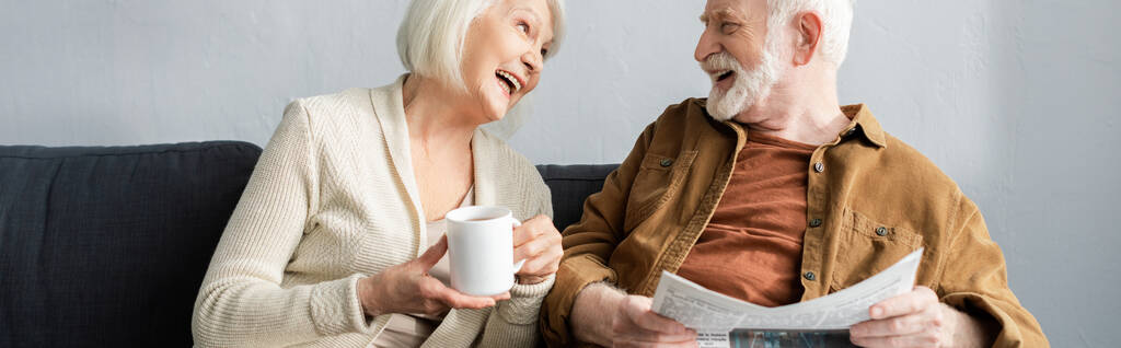 poziomy obraz starszej kobiety z filiżanką herbaty i jego męża z gazetą śmiejącą się podczas rozmowy na kanapie - Zdjęcie, obraz