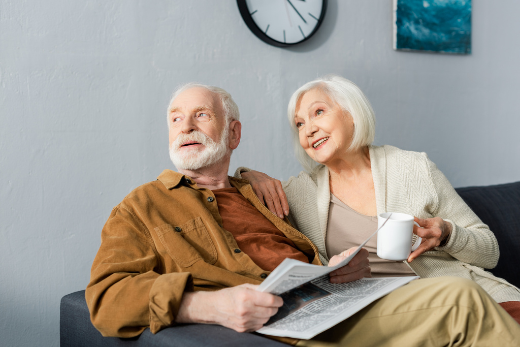 senior man with newspaper and his smiling wife with cup of tea looking away together - Photo, Image