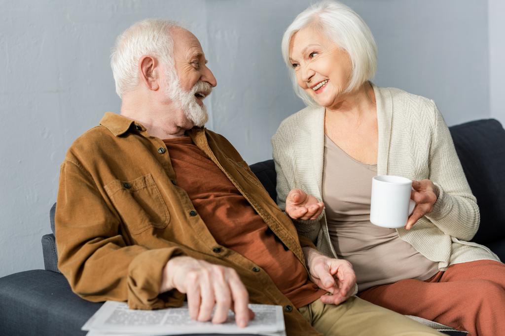 Gülümseyen kocasıyla konuşurken elinde bir fincan çay tutan neşeli yaşlı kadın. - Fotoğraf, Görsel