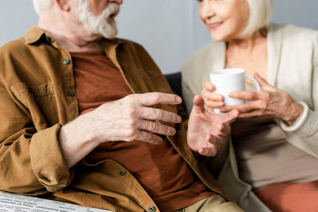 Selektiver Fokus eines älteren Mannes, der gestikuliert, während er mit seiner lächelnden Frau spricht, die eine Tasse Tee hält - Foto, Bild