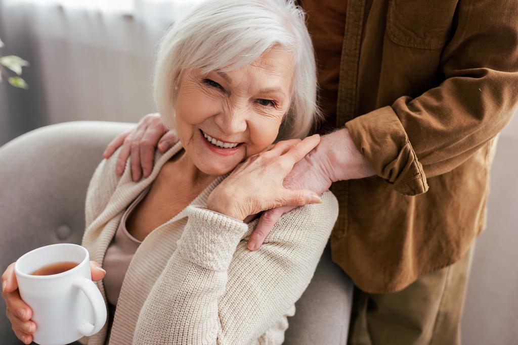 обрезанный вид мужчины трогательные плечи веселой старшей жены держа чашку чая и улыбаясь в камеру
 - Фото, изображение