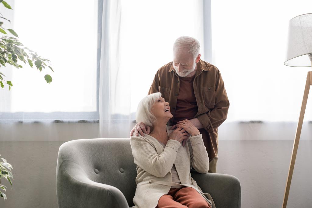 älterer Mann hält Hand und berührt Schulter der glücklichen Frau im Sessel - Foto, Bild