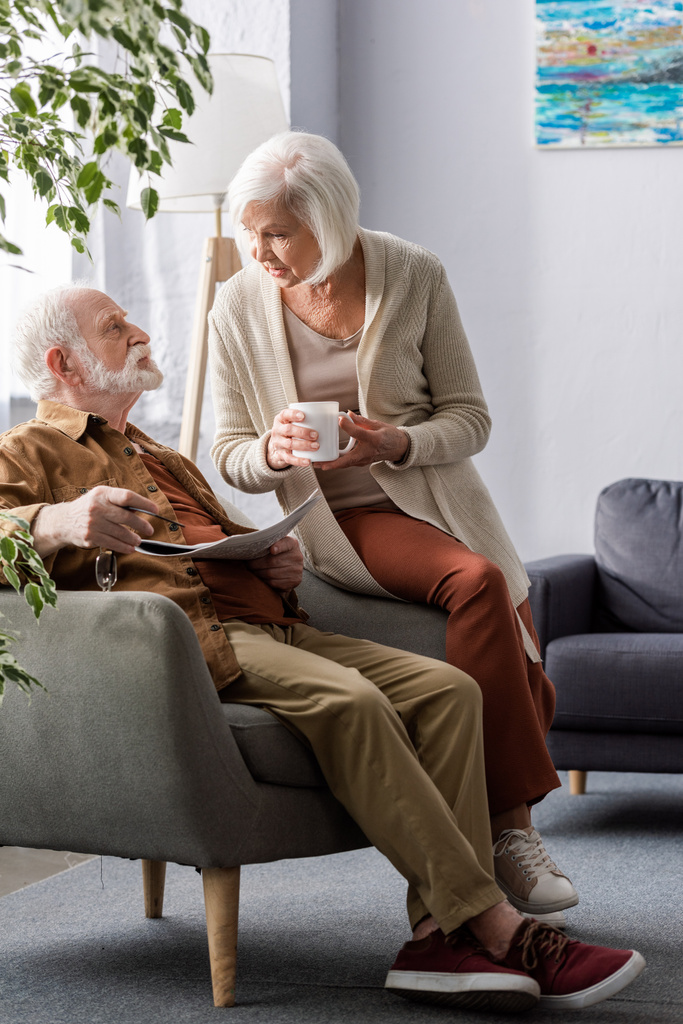 idős nő kezében csésze teát, miközben beszél férjjel ül karosszékben újságpapírral - Fotó, kép