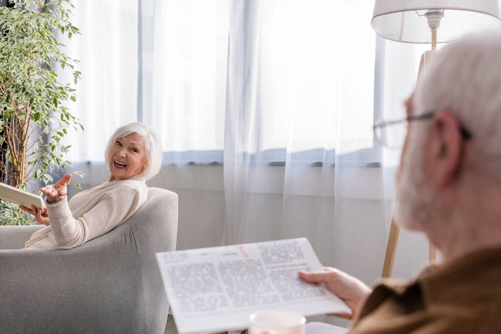 boldog nő ül karosszékben könyvvel, miközben beszél a férjével újság - Fotó, kép