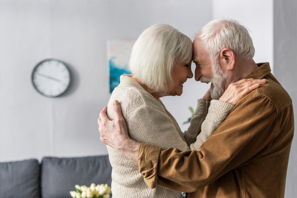 vista lateral de la feliz pareja de ancianos abrazando y riendo mientras están de pie cara a cara
 - Foto, imagen