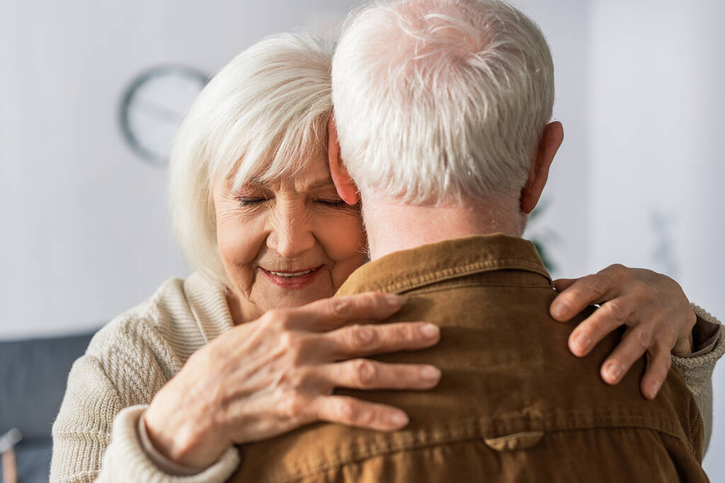 Rückansicht eines älteren Mannes, der von seiner glücklichen Frau mit geschlossenen Augen umarmt wird - Foto, Bild