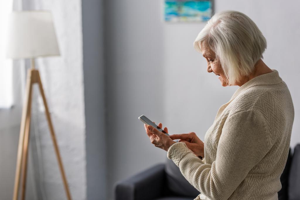 side view of smiling senior woman using smartphone at home - Photo, Image