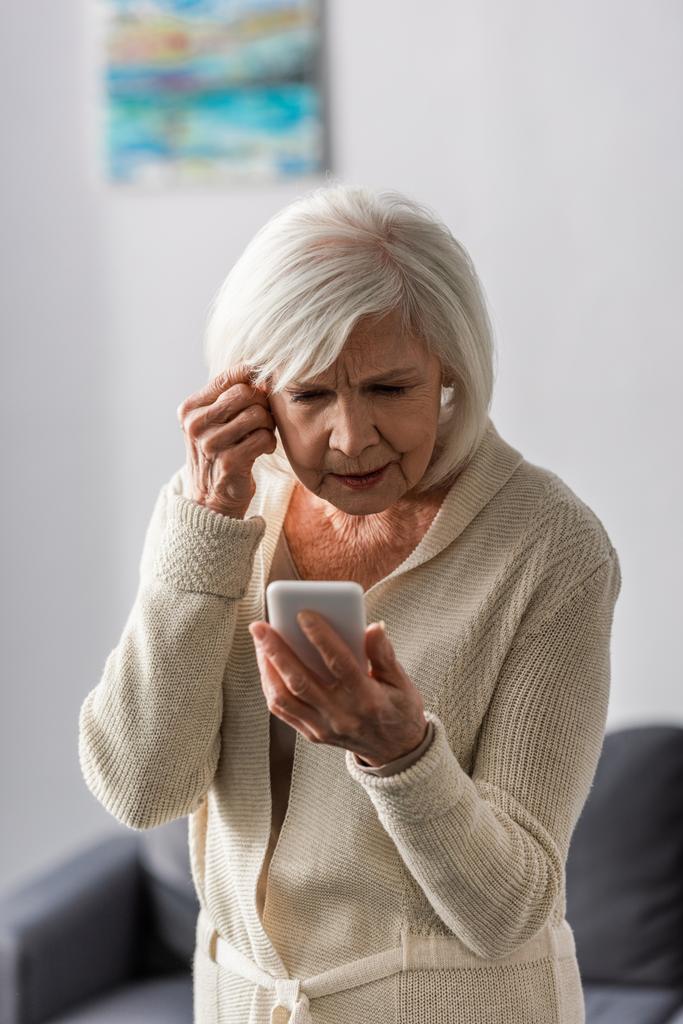 στοχαστική ηλικιωμένη γυναίκα που αγγίζει το κεφάλι ενώ χρησιμοποιεί το smartphone στο σπίτι - Φωτογραφία, εικόνα