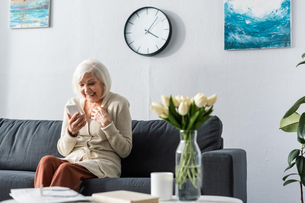 взволнованная пожилая женщина улыбается при использовании смартфона рядом со столом с тюльпанами букет
 - Фото, изображение