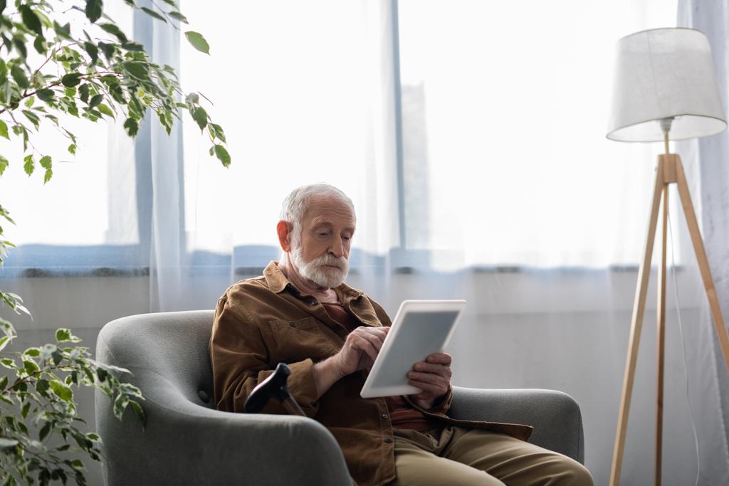 homme âgé concentré utilisant une tablette numérique tout en étant assis dans le fauteuil - Photo, image