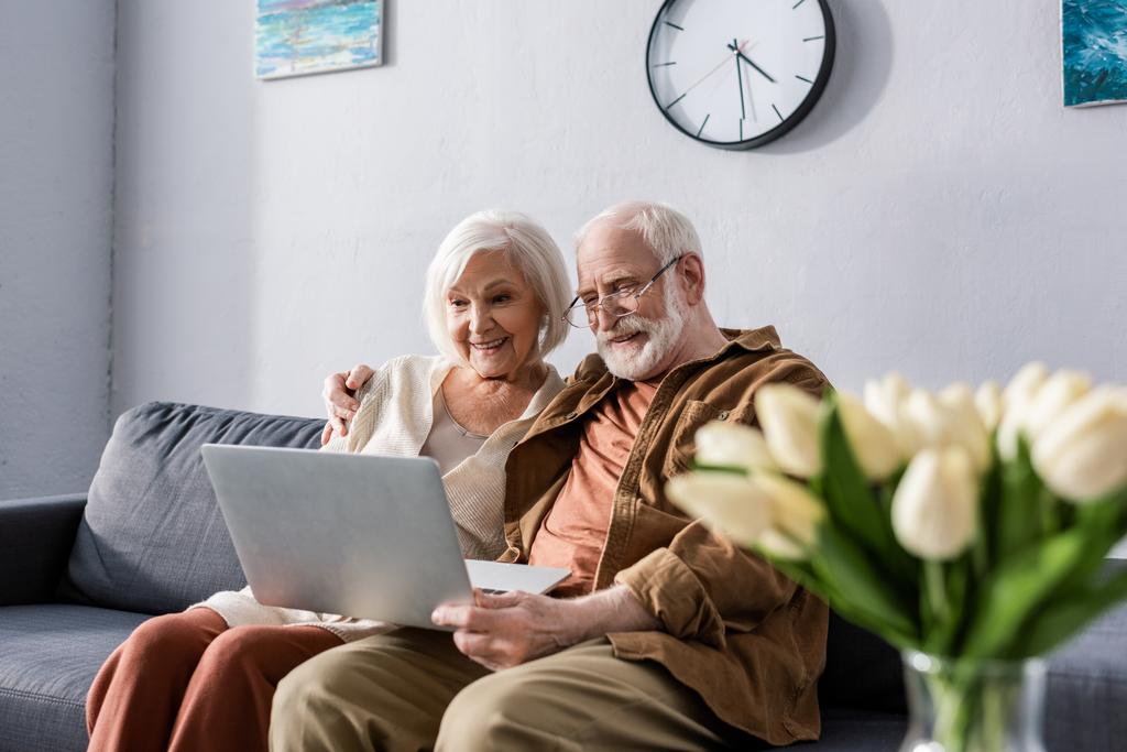 selectieve focus van gelukkig senior paar zitten op de bank en met behulp van laptop in de buurt boeket van tulpen - Foto, afbeelding