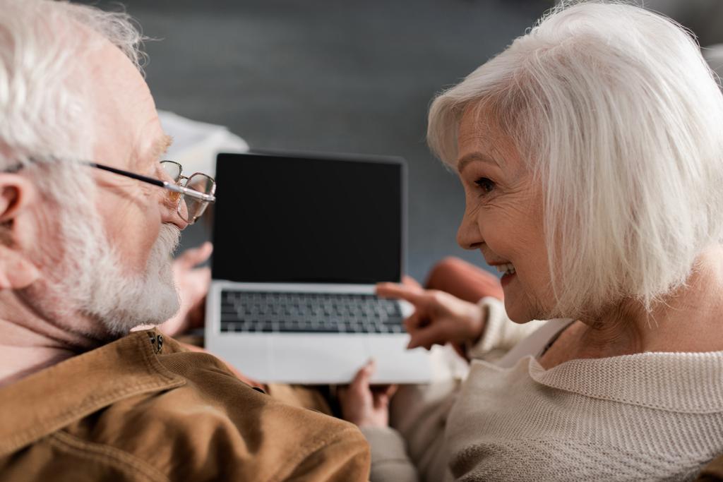 γενικά άποψη του χαμογελαστού ηλικιωμένη γυναίκα δείχνει στο φορητό υπολογιστή με λευκή οθόνη, ενώ κάθεται κοντά στο σύζυγο, επιλεκτική εστίαση - Φωτογραφία, εικόνα