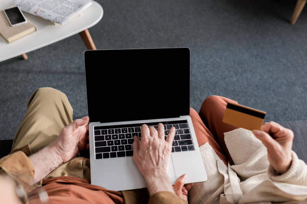 vista parcial del hombre mayor usando el ordenador portátil con la pantalla en blanco cerca de la esposa que sostiene la tarjeta de crédito, vista superior
 - Foto, Imagen