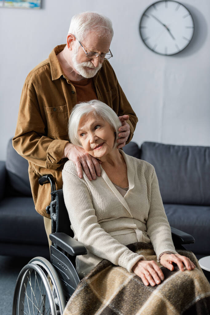 χαμογελαστή, ονειρεμένη ηλικιωμένη γυναίκα που κάθεται σε αναπηρική καρέκλα ενώ ο σύζυγος κρατά το χέρι στον ώμο της και αγγίζει τα μαλλιά της - Φωτογραφία, εικόνα