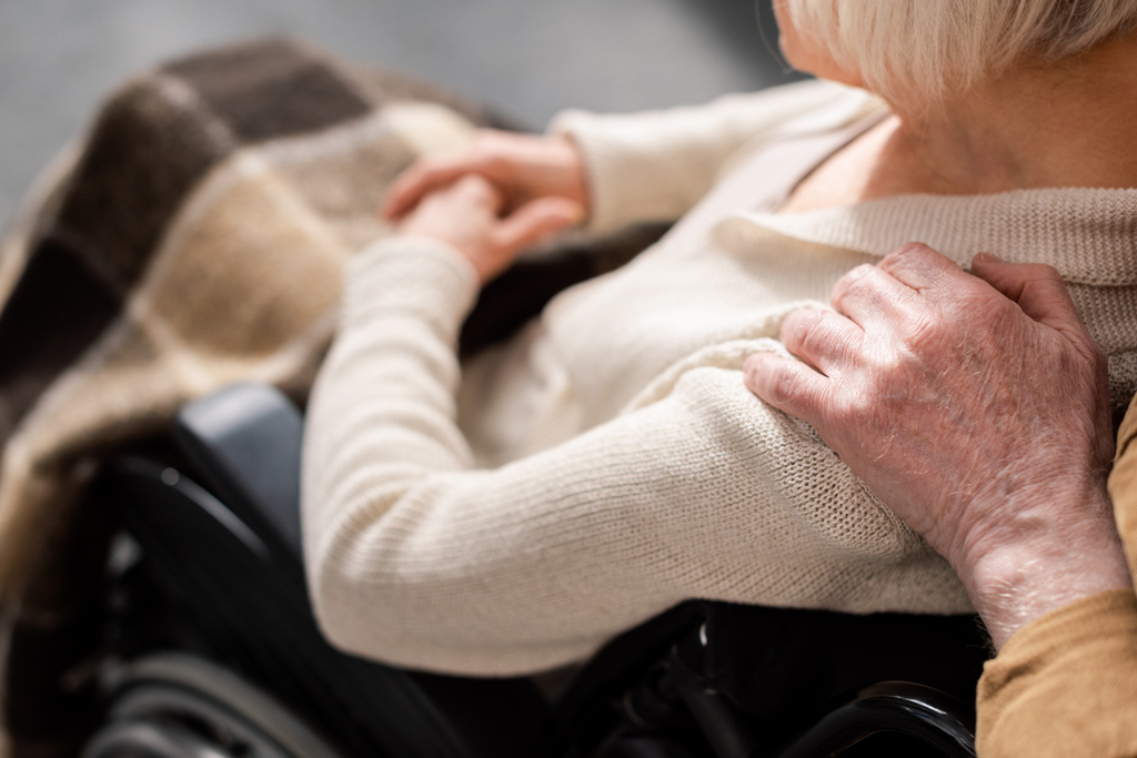 oříznutý pohled na muže dotýkajícího se ramene postižené, nemocné ženy sedící na invalidním vozíku - Fotografie, Obrázek