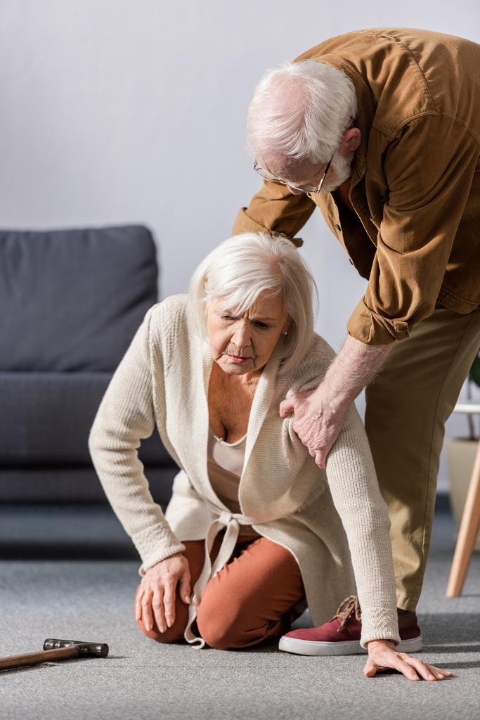 Пожилой мужчина помогает подняться упавшей жене, сидящей на полу
 - Фото, изображение