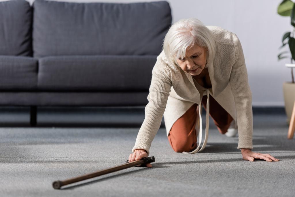 πεσμένη, αβοήθητη ηλικιωμένη γυναίκα που προσπαθεί να πάρει το μπαστούνι στο πάτωμα  - Φωτογραφία, εικόνα