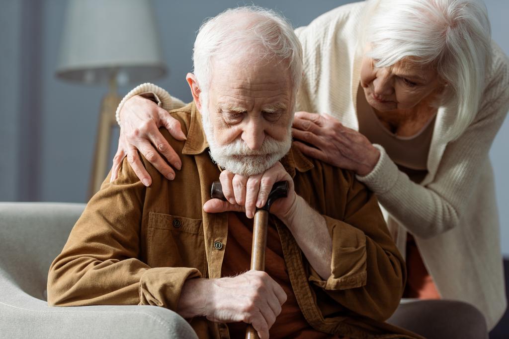 senior man, sick on dementia, sitting with walking stick, while wife looking at him and touching his shoulders - Photo, Image