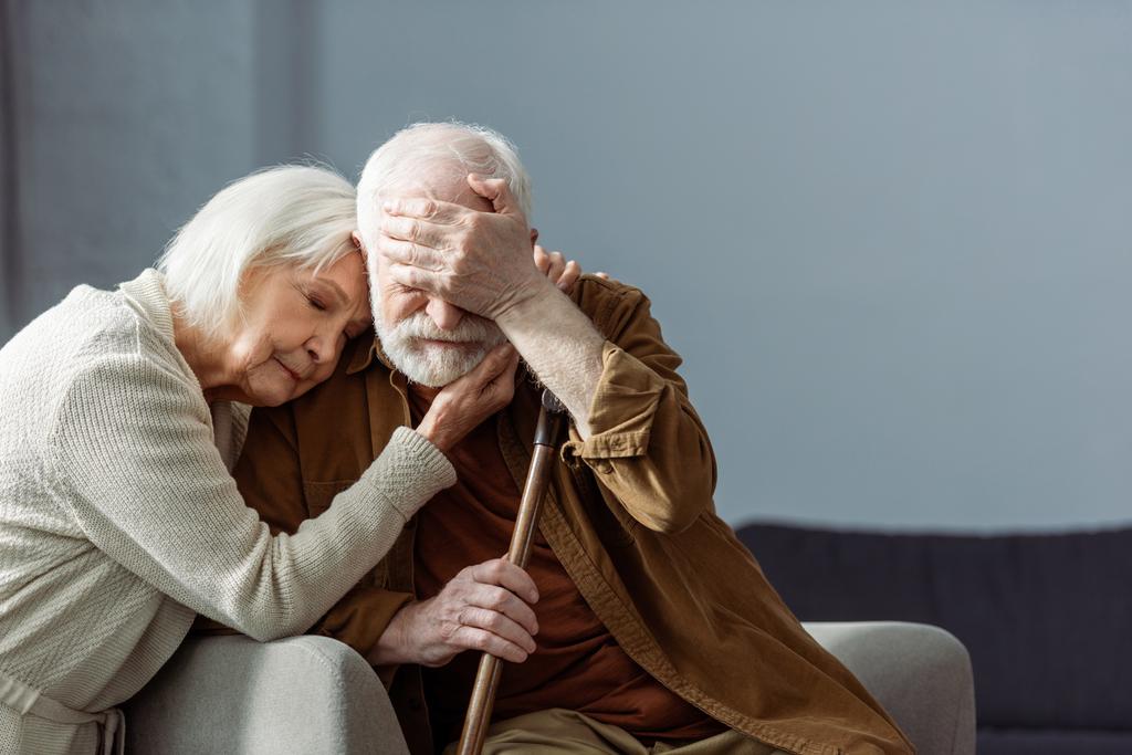 älterer Mann, an Demenz erkrankt, die Augen mit der Hand bedeckt, während seine Frau ihn mit geschlossenen Augen umarmt - Foto, Bild