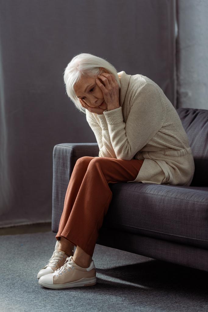 καταθλιπτική, μοναχική ηλικιωμένη γυναίκα που κρατιέται χέρι-χέρι ενώ κάθεται στον καναπέ - Φωτογραφία, εικόνα