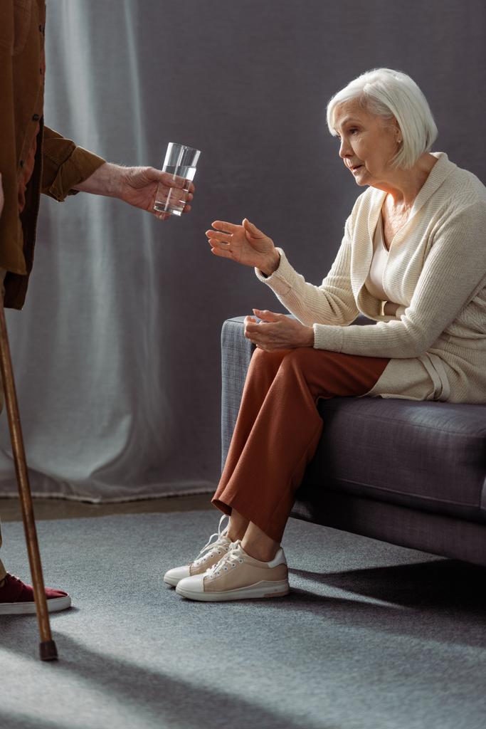 частковий вид на старшого чоловіка, який дає склянку води дружині, відчуває себе погано і сидить на дивані
 - Фото, зображення