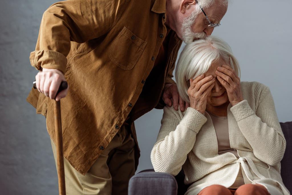 senior man with walking stick kissing head of diseased wife crying and covering eyes with hands - Photo, Image