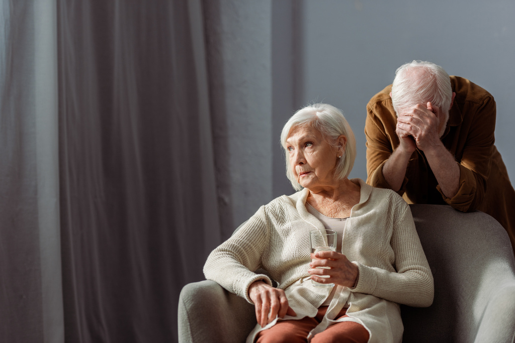 зневірений старший чоловік затемнює обличчя рукою, стоячи за хворою дружиною, тримаючи склянку води
 - Фото, зображення
