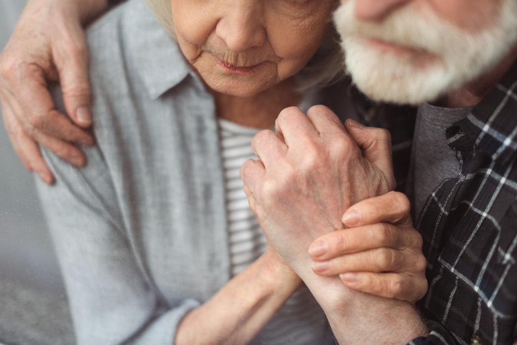 частичный вид пожилого мужчины и его жены, больного слабоумием, держащегося за руки, стоя у окна
 - Фото, изображение