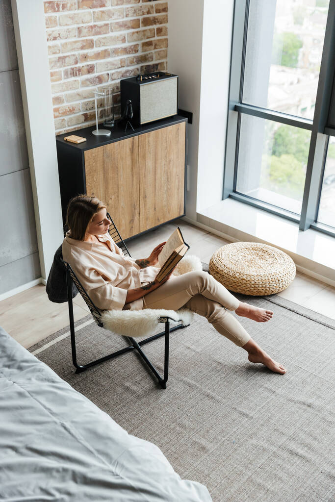 Imagen de una mujer joven pensando agradable en pijamas elegantes leyendo libro mientras está sentada en un sillón en la sala de estar
 - Foto, Imagen