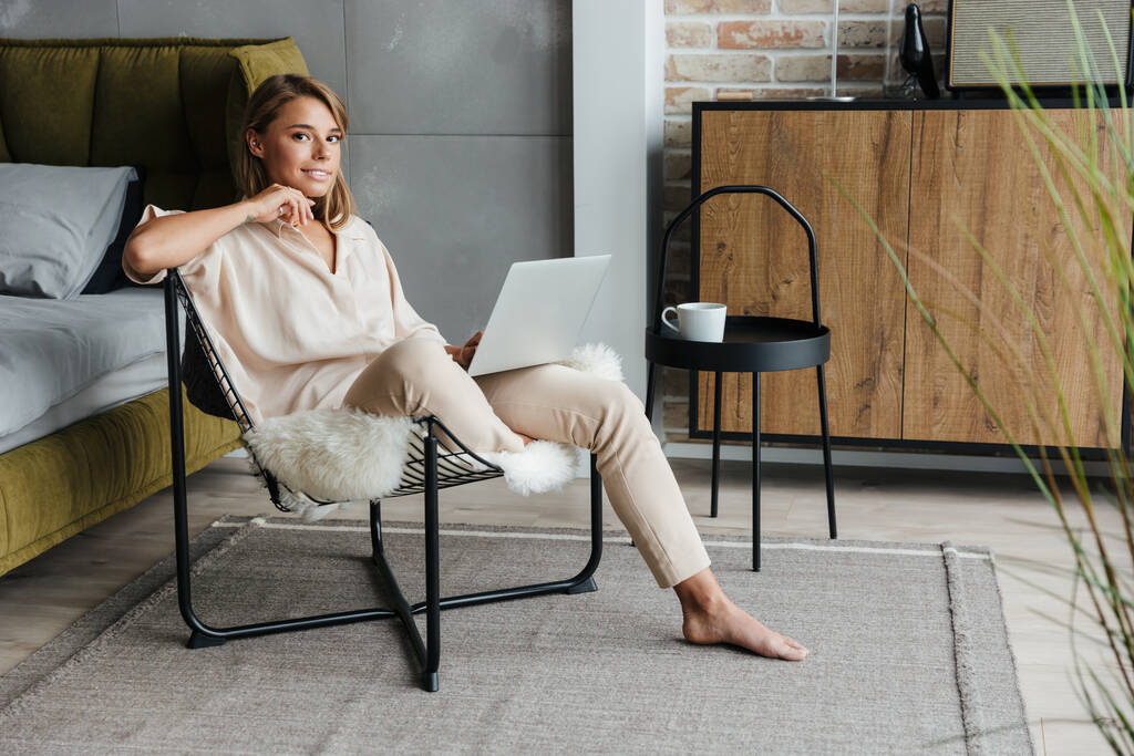Obrázek radostné blondýny pěkné ženy ve stylovém pyžamu pracující s laptopem, zatímco sedí na křesle v obývacím pokoji - Fotografie, Obrázek