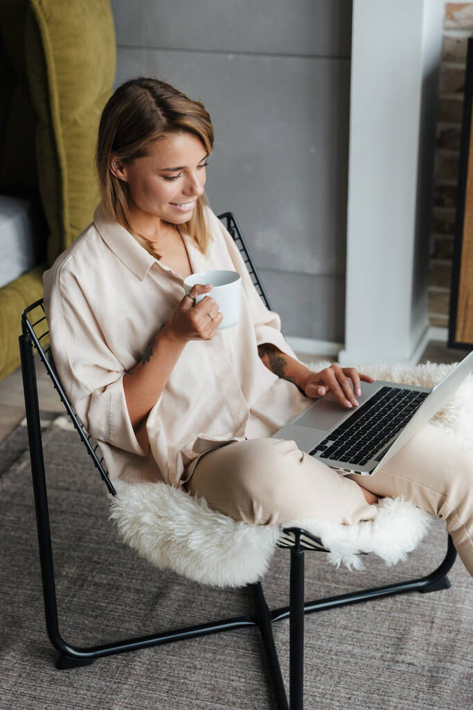 Изображение сосредоточенной милой женщины в стильной пижаме, пьющей чай и пользующейся ноутбуком, сидящей на кресле в гостиной
 - Фото, изображение